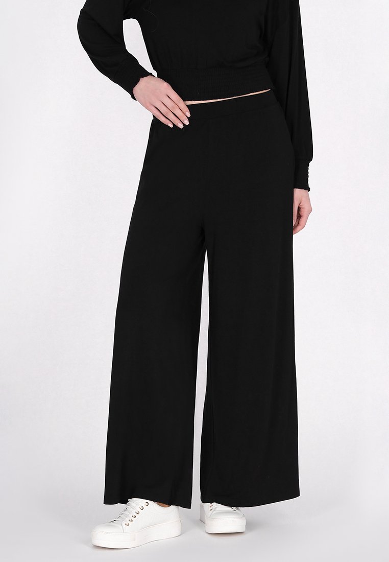 Schwarze weite Hosen aus weichem Stoff mit einem hohen elastischen Bund, kombiniert mit weißen Schuhen. Schlichtes Design, keine Muster.