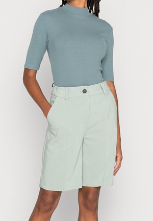 Femme portant un haut côtelé à manches courtes bleu sarcelle clair, rentré dans un short taille haute vert clair, coupé sur mesure, longueur genou.