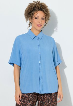 Vrouw met krullend haar, die een losse lichtblauwe blouse met knopen en bruine broek met luipaardprint draagt, staand tegen een witte achtergrond.