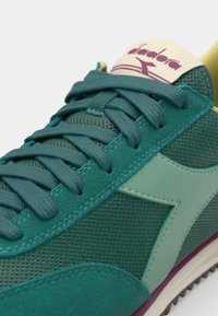 Teal sportieve schoen van mesh en suède materiaal, met gestructureerde veters, een contrasterende gele kraag en een paarse zool.