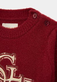 Maglione lavorato a maglia rosso con finitura testurizzata, scollatura rotonda e due bottoni scuri sulla spalla. Include un logo decorativo beige.