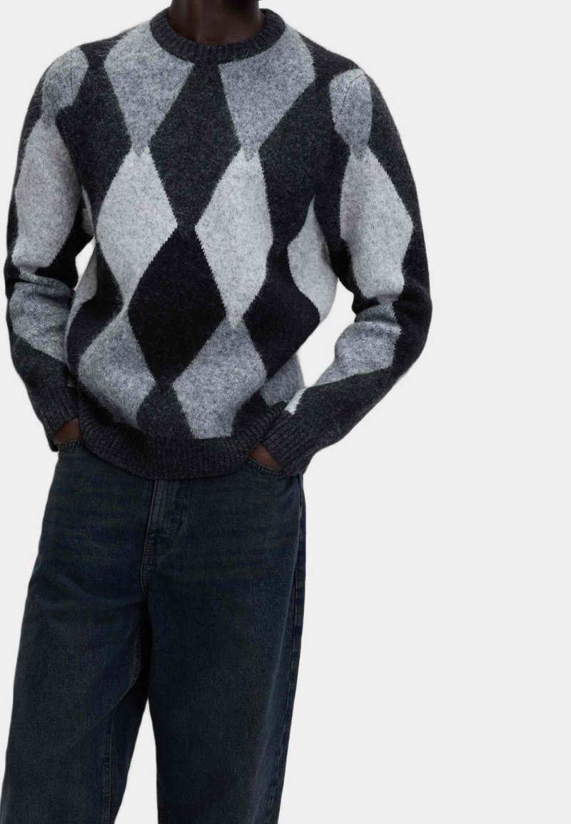 Personne portant un pull gris et noir duveteux à motif losange avec un jean foncé, mains dans les poches, vue des épaules aux genoux.