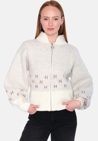 Strikket jakke med glidelås i kremfarge med ribbet kant og mansjetter. Har mønstrede detaljer med bokstavene "H" i lysbrun. Koselig og strukturert design.