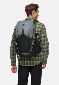 Zaino grigio e nero con superficie texturizzata, spallacci regolabili e tasche laterali per l'attrezzatura, indossato da una persona in abbigliamento a quadri.