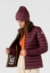 Chaqueta acolchada burdeos con forro amarillo, que presenta un estampado gráfico en el interior y un cuello alto. Combinada con una camiseta naranja y un gorro de canalé marrón granate.