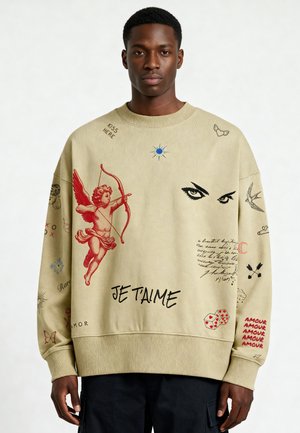 Homme portant un sweat-shirt beige avec un cupidon rouge, des yeux noirs, du texte manuscrit, des dés, des oiseaux, et des phrases comme "JE T'AIME" et "AMOUR" en rouge.