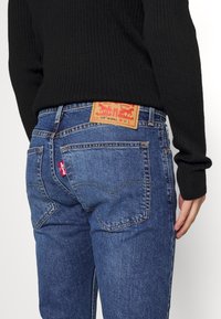 Blå denimbyxor med blekt tvätt, hög midja och kontrasterande orange sömmar. Har en röd Levi's-flik och en brun läderlogg.