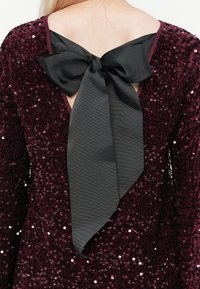 Rochie burgundă cu paiete, având un decolteu adânc în formă de V la spate și o funda mare din satin negru legată la gât, creând un contrast texturat.