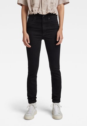 Jeans skinny noirs à taille haute, avec des passants de ceinture, un bouton à l'avant et une texture lisse. Portés avec des sneakers blanches.