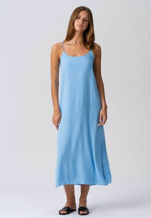 Femme debout, portant une robe maxi bleu clair sans manches avec de fines bretelles et des sandales noires à enfiler, devant un fond clair uni.