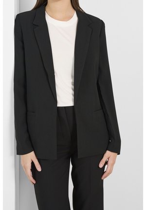 Mujer que lleva un blazer negro y pantalones con una camiseta blanca de cuello redondo, de pie frente a una pared lisa de color claro.