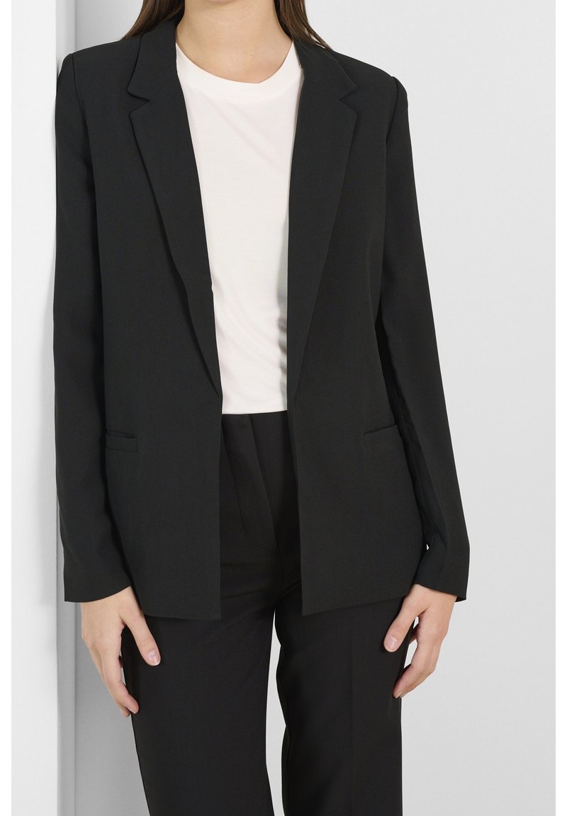 Femme portant un blazer noir et un pantalon avec un t-shirt blanc à col rond, debout contre un mur uni de couleur claire.
