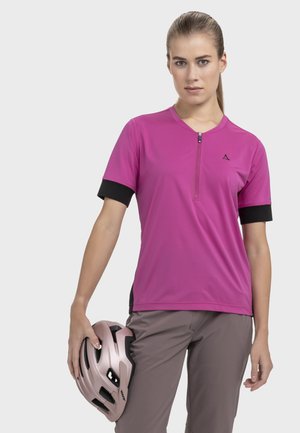 Frau trägt ein pinkfarbenes kurzärmeliges Radsporttrikot und graue Hosen, hält ihren metallisch pinkfarbenen Fahrradhelm in der linken Hand.