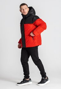 Rød og sort puffjakke med lynlåslukning, hætte og sidelommer. Jakken har et farveblokdesign og en quiltet tekstur.
