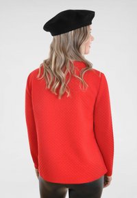 Rode gestructureerde trui met lange mouwen, een gebogen zoom en een zwarte baret. De stof lijkt zacht en gemusterd, geschikt voor casual dragen.