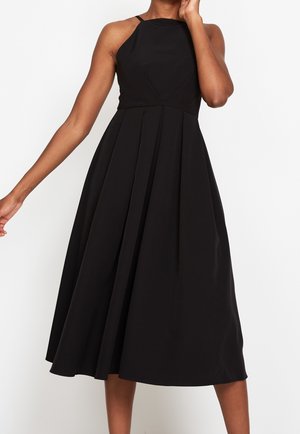Kobieta ubrana w czarną, bezrękawnikową sukienkę midi z dopasowanym gorsetem i rozkloszowaną plisowaną spódnicą, stojąca na tle jednolitego białego tła.