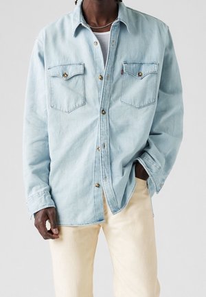 Mand iført lyseblå denim skjorte med knapper over hvid tanktop og beige bukser med den ene hånd i lommen mod ensfarvet baggrund.