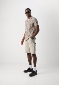 Camisa de manga curta com padrão em castanho e bege, combinada com calções bege e sapatos pretos. Óculos de sol completam o visual.