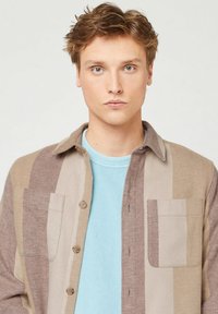 Camicia button-up con una combinazione di righe marroni e beige, realizzata in tessuto texturizzato e dotata di una tasca sul petto, indossata sopra una maglietta a coste di colore azzurro chiaro.