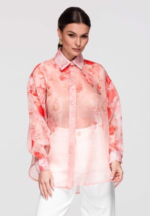 Vrouw met donker haar draagt een doorschijnende roze bloemen oversized blouse en witte broek, staand tegen een effen lichte achtergrond.