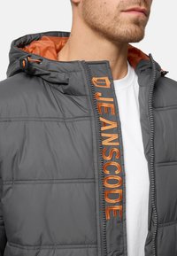 Homme portant une doudoune grise avec une doublure orange et "JEANSCODE" brodé en orange le long de l'ouverture de la fermeture éclair.