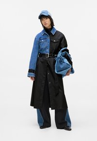 Jeansowa i czarna tkaninowa kurtka z pasem w talii, długimi rękawami oraz mieszanką tekstur. Pasująca niebieska czapka i niebieska torba na sznurku.