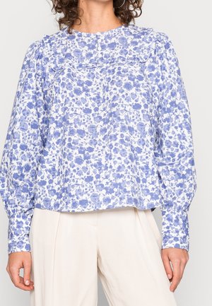 Kvinde iført en langærmet hvid bluse med blåt blomsterprint, puffede ærmer og tre hvide knapper nær halsudskæringen, kombineret med beige bukser.