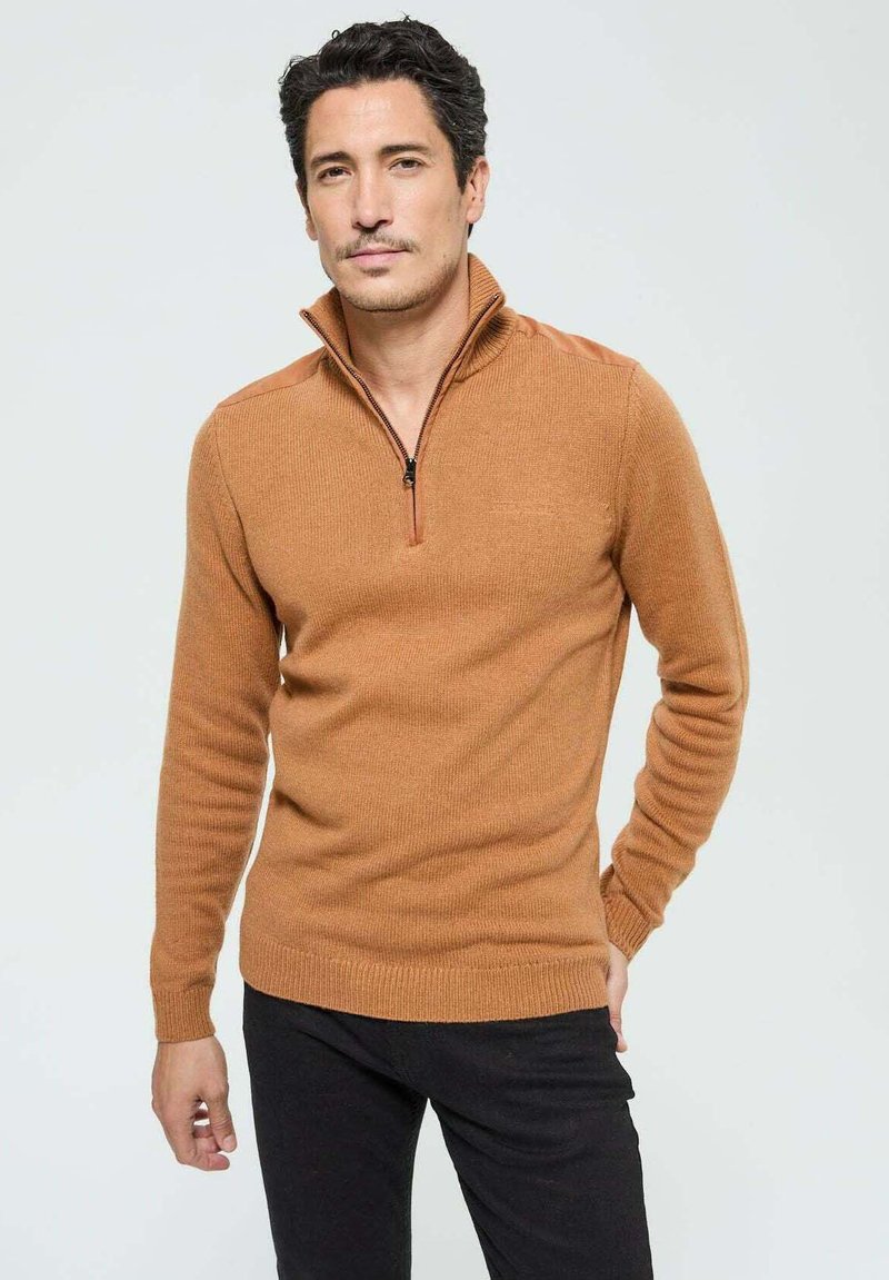 Pull à col zippé marron avec texture côtelée et accents d'épaules contrastés. Manches longues et design ajusté, associé à un pantalon sombre.