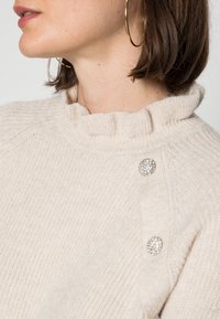 Pull en tricot beige clair avec un col montant, présentant une texture côtelée et des boutons décoratifs en métal argenté sur l'épaule.