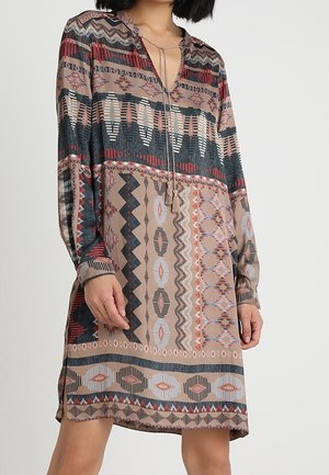 Vrouw draagt een jurk met lange mouwen tot op de knie met meerkleurige geometrische en tribale patronen, inclusief kwastdetail bij de halslijn.