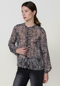 Fekete-fehér paisley mintás blúz gombos elülső résszel, hosszú ujjakkal és átlátszó anyagból, sötét farmernadrág fölé öltözve.