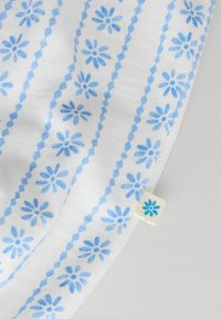 Tessuto bianco con un motivo ripetuto di piccoli fiori blu e linee tratteggiate; include un piccolo cartellino bianco con un accento floreale blu.