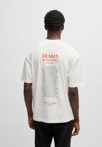 T-shirt di cotone color crema con maniche corte, con testo rosso e arancione sul retro, che mostra le date e le località del tour.