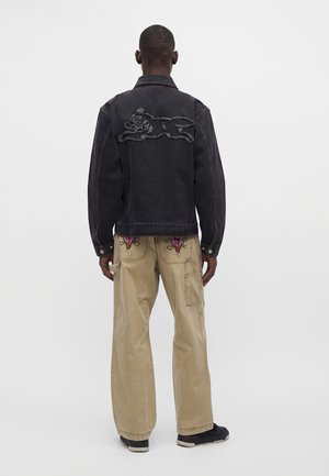 Homme portant une veste en jean noire avec un motif tigre dans le dos et un pantalon beige avec des crânes brodés colorés sur les poches arrière.