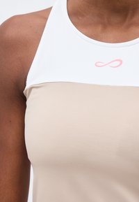 Gros plan d'une personne portant un haut beige et blanc sans manches avec un symbole infini rose près de l'encolure.