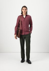 Chemise boutonnière bordeaux à manches longues, boutons foncés. Pantalon vert olive, texture lisse. Sac à bandoulière marron, sangle réglable. Chaussures noires.