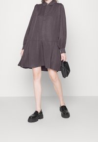 Abito grigio scuro con colletto, maniche plissettate e orlo svasato. La modella tiene una piccola borsa nera e indossa scarpe nere lucide.