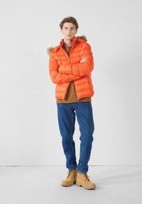 Junger Mann steht mit verschränkten Armen, trägt eine orange Daunenjacke, ein braunes Hemd, blaue Jeans und beige Schnürstiefel vor einer weißen Wand.