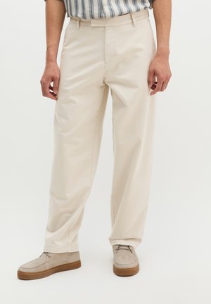 Man draagt beige broek met rechte pijpen, lichtbruine suède schoenen en een gestreept ingestopt overhemd, staand tegen een effen achtergrond.