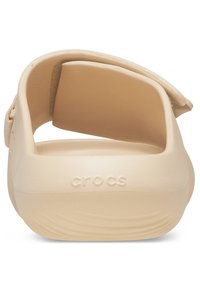 Beige Crocs sandaal, gemaakt van zacht rubbermateriaal, met een afgeronde achterkant en een getextureerde voetbed. Beschikt over een verstelbare band en een lage hak.
