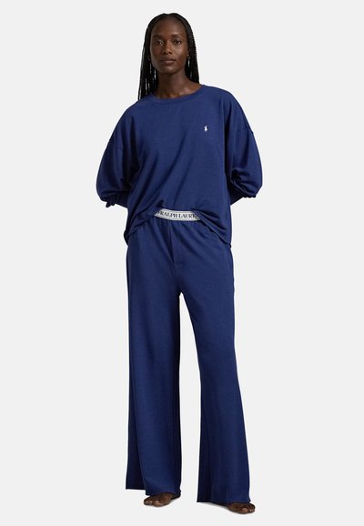 Γυναίκα που στέκεται ξυπόλητη, φοράει άνετο, χαλαρό μπλε σκούρο φούτερ και αντίστοιχο φαρδύ παντελόνι με εμφανές λάστιχο μέσης Ralph Lauren.
