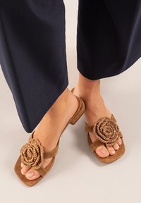 Sandalias de ante marrón con suelas planas, que presentan un acento de flor trenzada en la correa del dedo del pie. El diseño es abierto y casual.