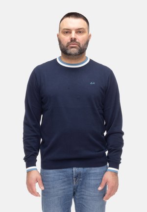 Uomo con capelli corti scuri e barba, che indossa un maglione blu navy con colletto e polsini a righe bianche e blu, e jeans azzurri, in piedi.