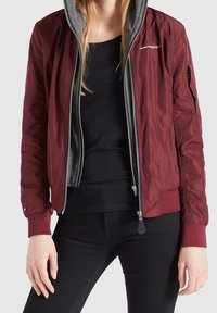 Vínově červená bomber bunda se šedou kapucí, se zipem vpředu, žebrovanými manžetami a bočními kapsami. Vyrobena z hladké, lehké látky.