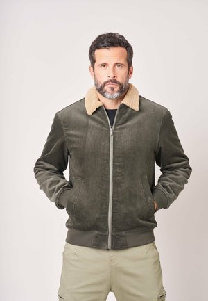 Man met baard draagt een groene corduroy jas met beige bontkraag en beige broek, staand met handen in de zakken tegen een effen achtergrond.