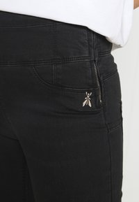 Vaqueros de mezclilla negra con un bolsillo con cremallera y un pequeño logotipo plateado de insecto como acento. Textura suave con detalles de costura estándar.