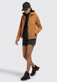 Tan waterproof jacket with a hood, front zip, and two chest pockets. Worn over a black top and denim shorts, paired with black sneakers.