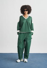 Πράσινο αθλητικό τοπ με λευκές λεπτομέρειες, V-λαιμόκοψη και μακριά μανίκια, σε συνδυασμό με χαλαρά πράσινα παντελόνια με λευκή ραφή και τσέπες.