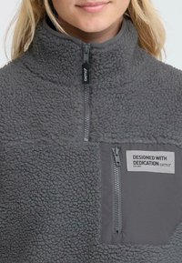 Hallides hall pullover kõrge kraega, tekstuuriga pind, neljandiku pikkuse tõmblukk ning eesmine tasku, millel on brändi silt ja tekst.