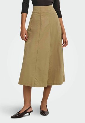 Žena má na sebe béžovú midi sukňu do A s vysokým pásom, čierny top a čierne lodičky slingback so špicatou špičkou.